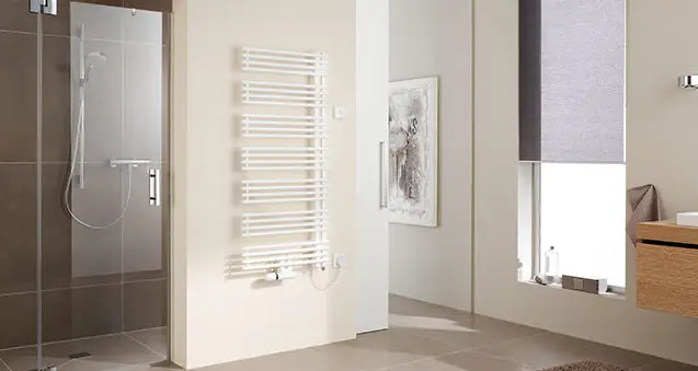 Weißer Kermi-Heizkörper mit Längsrippen an der Wand eines modernen Badezimmers, daneben eine Glasdusche und ein Fenster mit Rollladen.